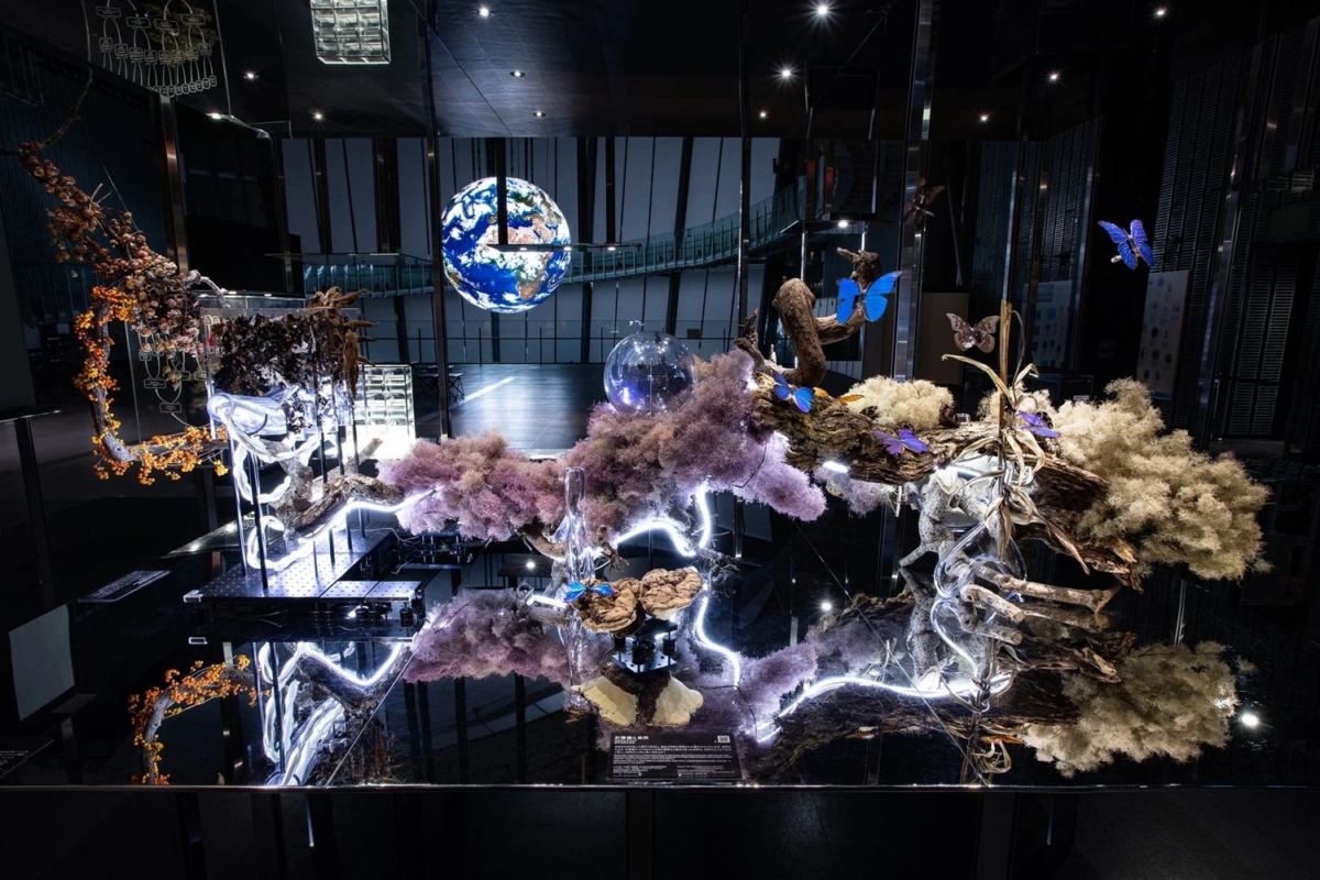 地球儀が浮かぶ暗い空間に、枯れ木や苔、青い蝶、白い光が絡み合う幻想的なインスタレーション。自然の循環と生命の輝きをテーマにした現代アート作品です。