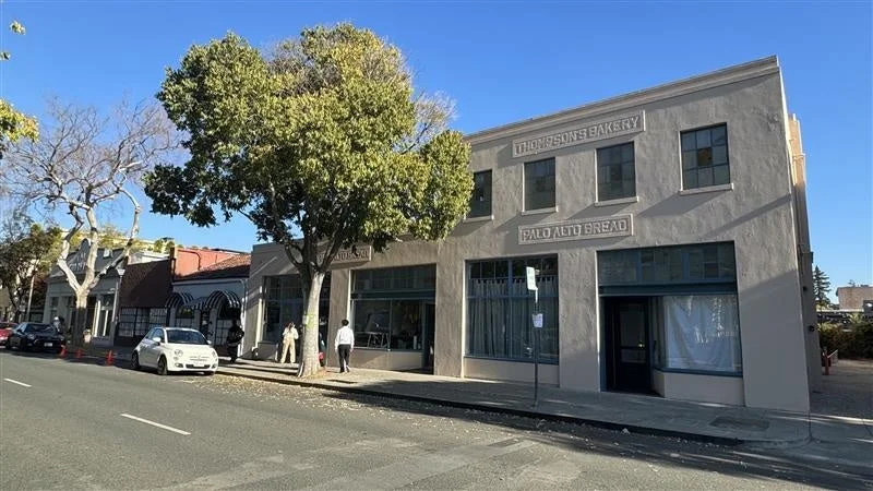 「THOMPSON'S BAKERY」と「PALO ALTO BREAD」の看板がある建物が写る街並みの写真です。青空の下、通りには白い車が停まり、数人の通行人が見えます。歴史を感じさせる建物と緑豊かな木々が特徴的な昼間の風景です。