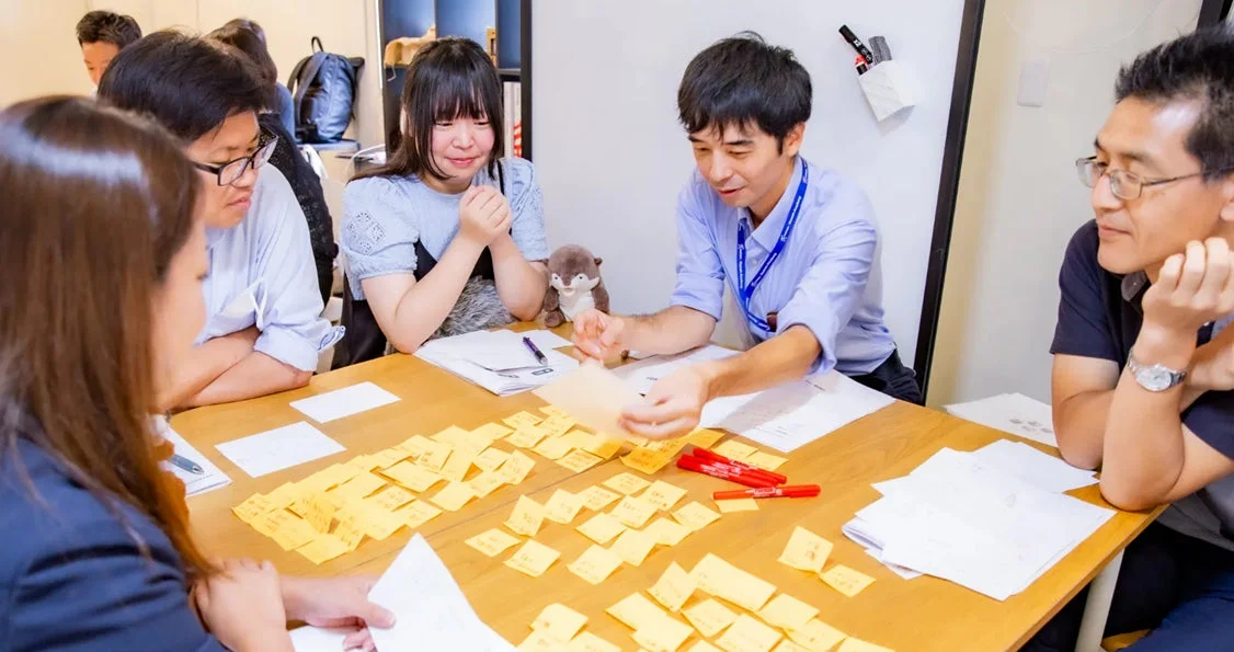 数人の人々がテーブルを囲み、ワークショップかブレインストーミングを行っている画像です。