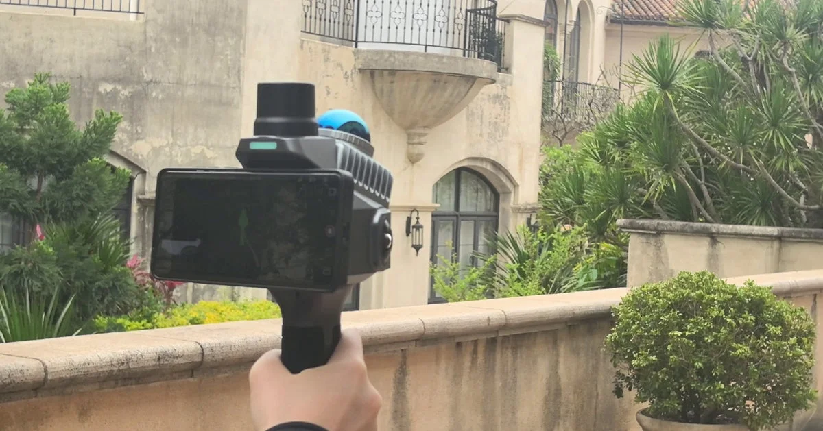 人物が手持ちの計測デバイスを構え、豪華な建物とその周囲の豊かな植栽を撮影している様子。デバイスの画面にはグラフィカルな表示が見られます。