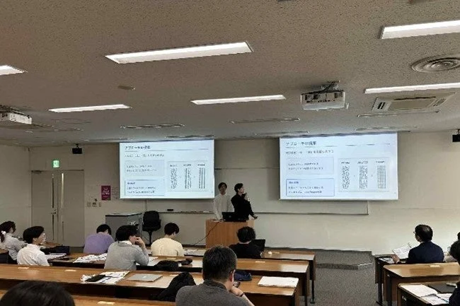 教室のような会場でプレゼンテーションが行われている様子。二人の発表者が壇上に立ち、二つのスクリーンには「アプローチの提案」と「アプローチの成果」と題されたスライドが表示されている。多くの聴講者が机に着席し、発表を聞いている。