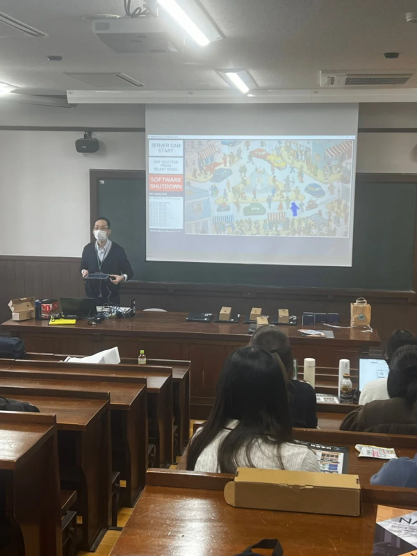 講堂で講師がプレゼンテーションを行っており、プロジェクターには賑やかな街のイラストと「SOFTWARE SHUTDOWN」などの文字が映し出されています。複数の学生が講義を聴講している様子がうかがえます。