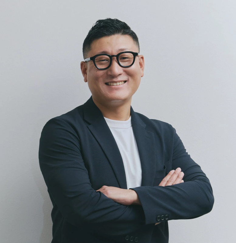 黒縁メガネをかけたアジア人男性が、黒いジャケットと白いTシャツを着用し、腕を組んで笑顔で写っています。