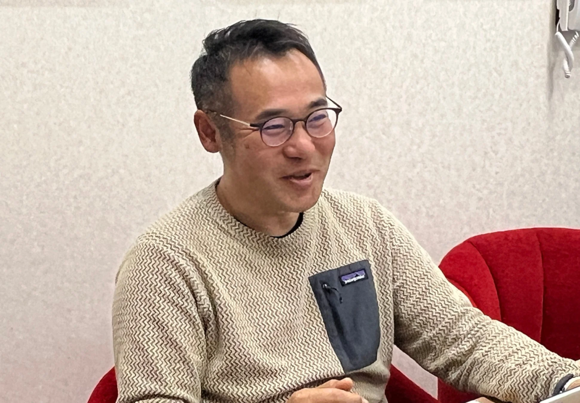 眼鏡をかけた男性が、模様入りのセーターを着て笑顔で座っているポートレート。室内でリラックスした雰囲気で会話している様子が伺える。