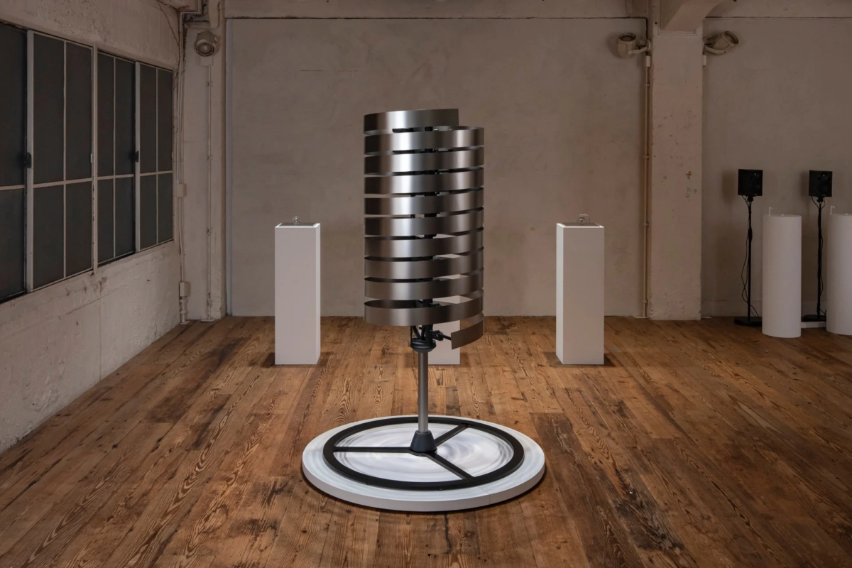 木製の床と白い壁の広々とした空間に、金属製の水平な帯が螺旋状に重なり合った現代アートのインスタレーションが展示されています。ミニマルなデザインの彫刻が中央に配置され、展示会場のような雰囲気を醸し出しています。