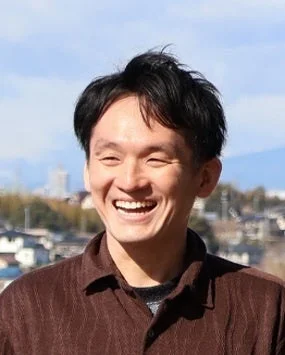 屋外で明るく笑う男性のポートレートです。男性は短く黒い髪で、焦げ茶色のシャツを着ています。背景には住宅街が広がり、晴れた空が見えます。