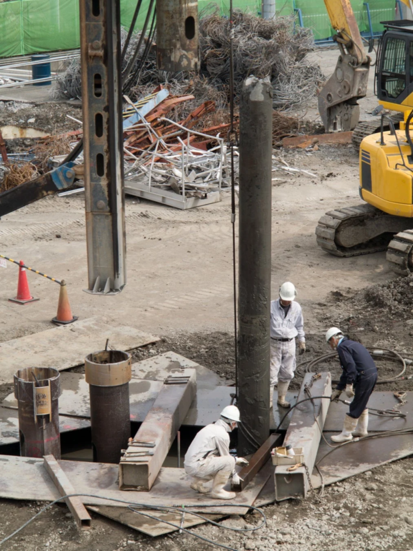 建設現場で数人の作業員が基礎工事や杭打ち作業を行っている様子を上空から捉えた画像です。周囲には重機や大量の廃材が積まれており、大規模な工事が進行中であることが伺えます。