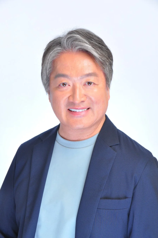 白背景の前で、紺色のジャケットと水色のTシャツを着用し、笑顔でカメラを見つめる白髪の中年男性のポートレートです。親しみやすい印象を与えます。