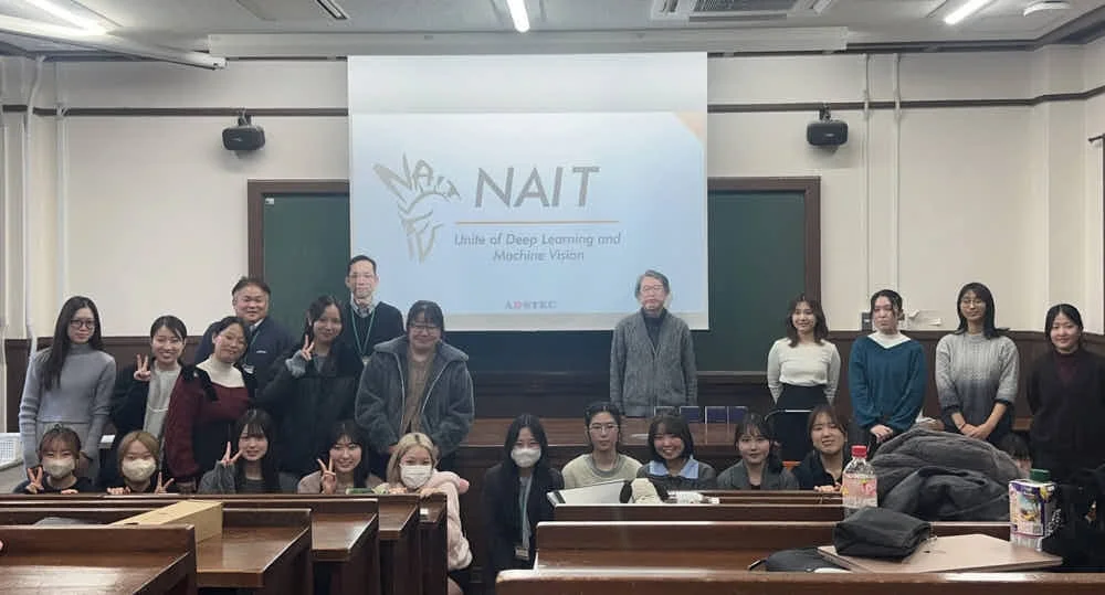 大学の教室で、ディープラーニングとマシンビジョンに関連するNAITのメンバーと思われる学生と教員が集合写真を撮っています。多くの人がピースサインをしています。
