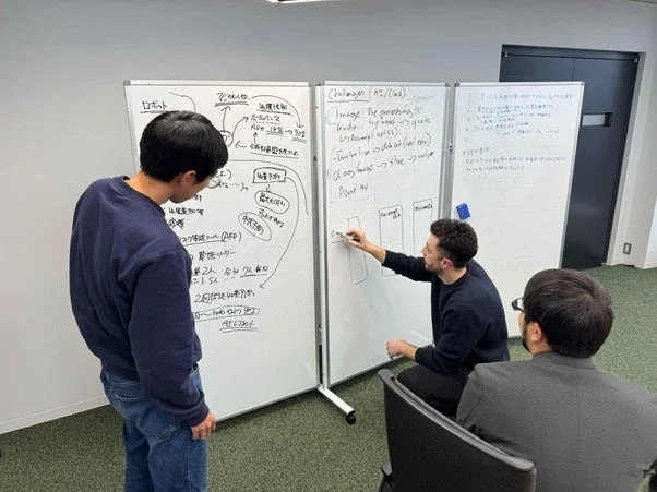 オフィスで3人の男性がホワイトボードを使って活発に議論している様子です。