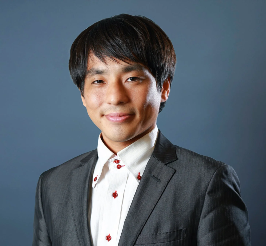 暗い背景の前で、ダークスーツと赤いボタンの白いシャツを着たアジア人男性のポートレート。男性はカメラに向かって微笑んでいます。
