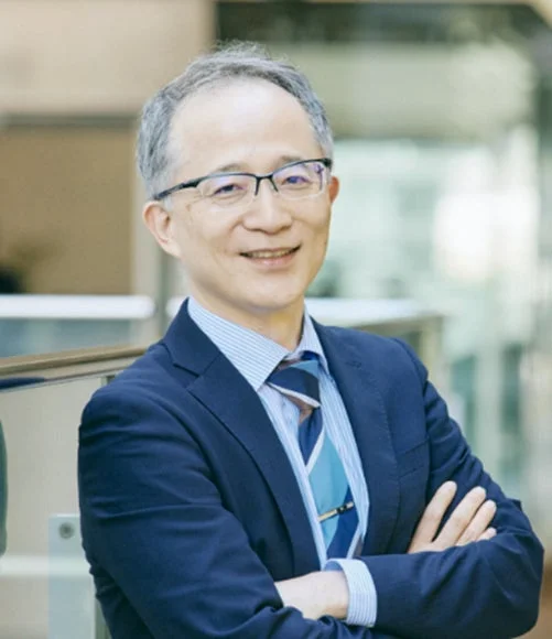 青いスーツとストライプのシャツ、ネクタイを着用し、眼鏡をかけたアジア系の男性が腕を組み、穏やかに微笑んでいる。プロフェッショナルな雰囲気のポートレート写真です。