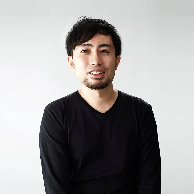 白い背景の前で、黒いVネックの長袖シャツを着た笑顔の男性のポートレートです。短髪と髭が特徴的で、親しみやすい印象を与えます。