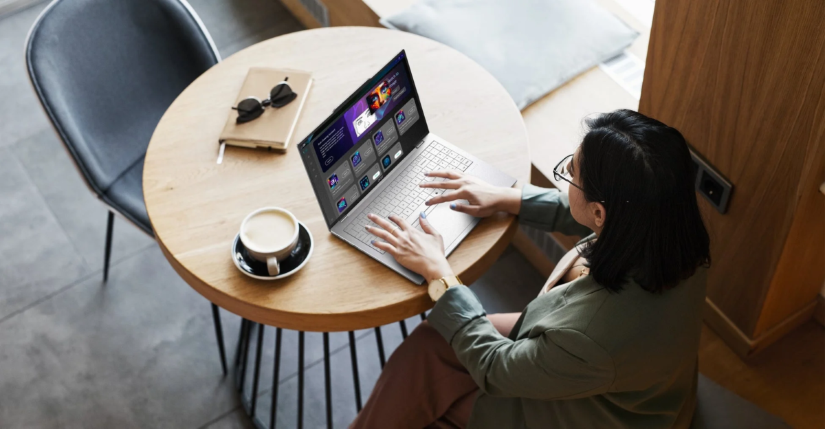 カフェで女性がノートパソコンを使って作業している様子。テーブルにはコーヒーとノートがあり、パソコン画面にはAI関連のアプリケーションが見える。