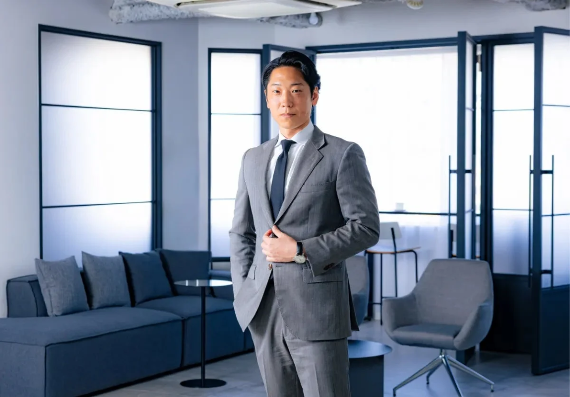 グレーのスーツを着た日本人男性が、モダンなオフィス空間で正面を向いて立っています。背景には大きな窓とソファ、椅子が見えます。
