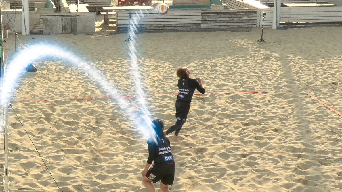 砂浜のコートでビーチバレーをする2人の選手が写っています。ボールが空中にあり、1人の選手がそれを見上げています。ボールの軌跡を示すような光のエフェクトが見られます。