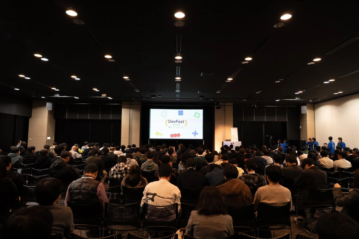 DevFest Tokyo 2025が開催されている会場で、多数の参加者がスクリーンに映し出されたロゴを見ている様子。