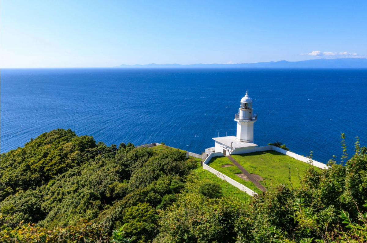 青い海と青い空が広がる岬に立つ白い灯台の風景