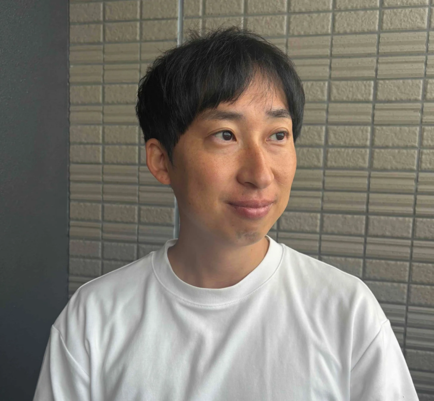 白いTシャツを着たアジア人男性のポートレート。男性は穏やかな表情で、画面の右方向を見ています。背景はタイル張りの壁です。