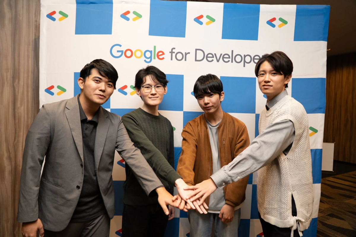 「Google for Developer」のバナーを背景に、4人の若い男性が手を重ねてポーズをとっています。