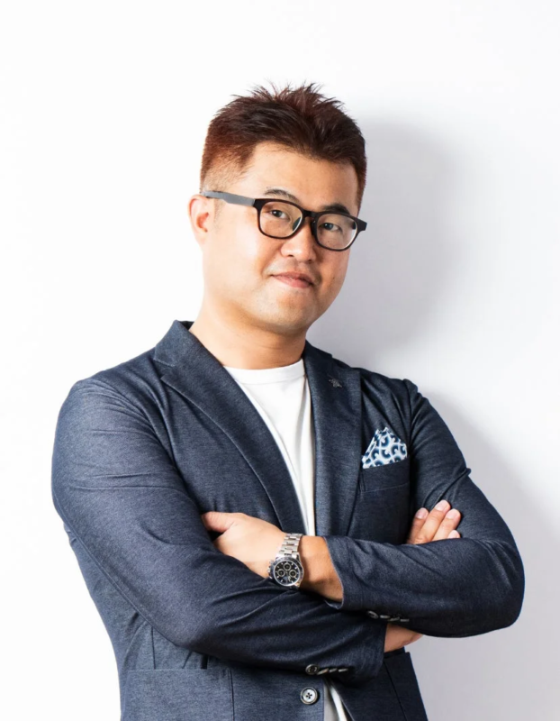 白い背景の前で、メガネをかけた日本人男性が紺色のブレザーと白Tシャツ姿で腕を組み、カメラ目線で微笑んでいます。