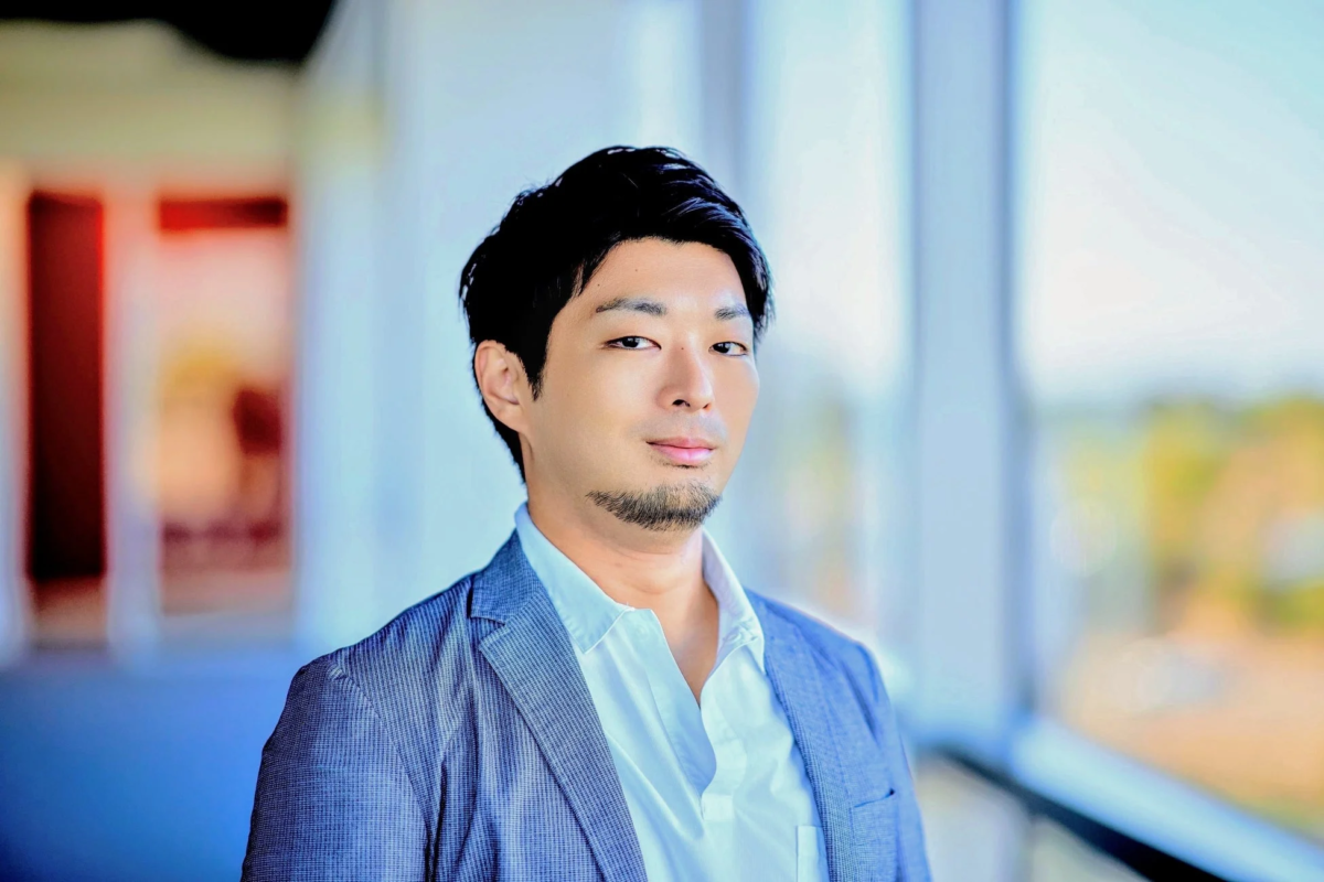 青いジャケットと白いシャツを着用し、髭を蓄えた日本人男性が、明るい窓のある屋内でカメラを真っ直ぐ見つめているポートレート写真