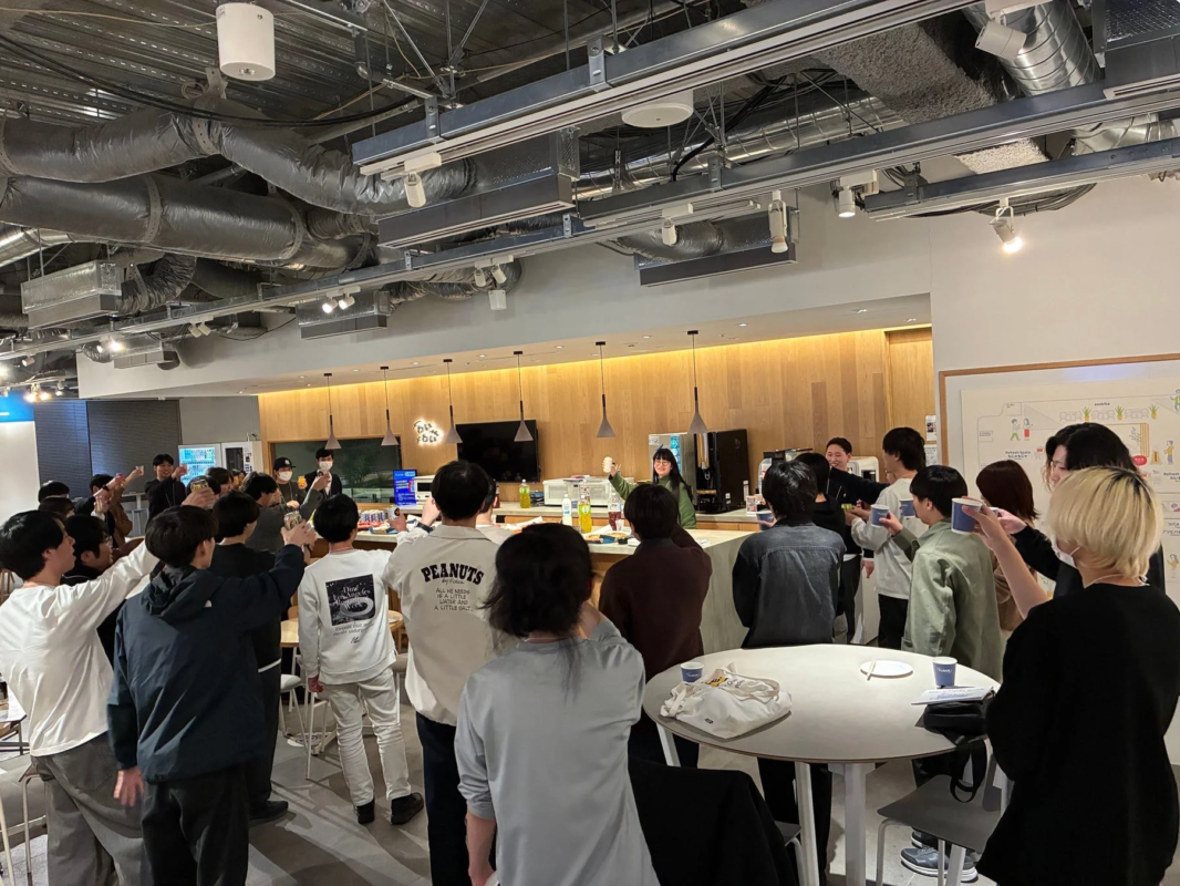 多くの人々がモダンなオフィススペースや共有エリアに集まり、飲み物を片手に交流している様子