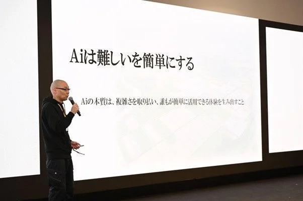 一人の男性がステージ上でマイクを手にプレゼンテーションを行っています。背景の大きなスクリーンには「Aiは難しいを簡単にする」というメインメッセージと、「Aiの本質は、複雑さを取り扱い、誰もが簡単に活用できる体験を生み出すこと」という説明文が表示されており、AIの役割について語られている場面です。