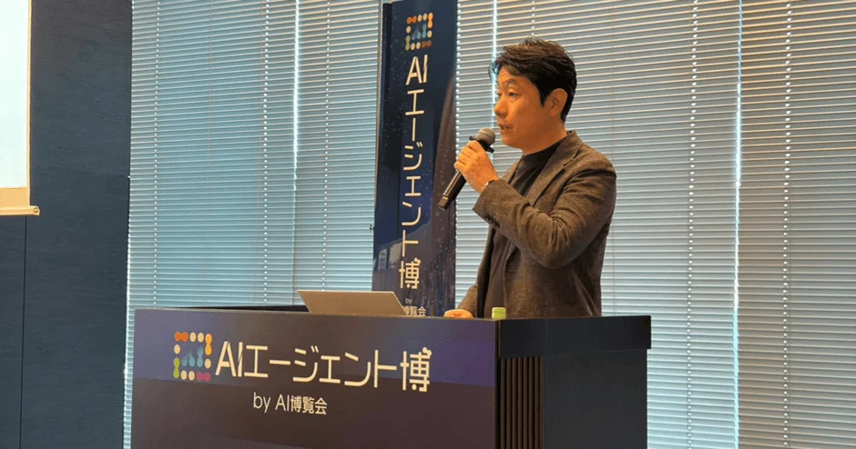 男性が「AIエージェント博」と書かれた壇上でマイクを持って講演している様子