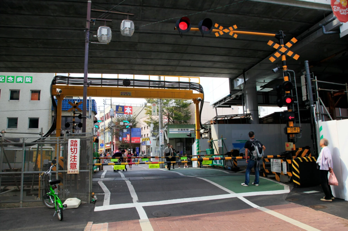 高架下にある日本の踏切の風景。赤信号と遮断機が下り、人々が電車を待っている様子