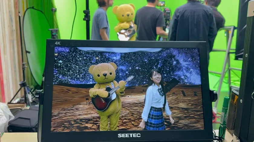撮影スタジオでグリーンバックを活用した映像制作の様子。モニターには、宇宙と砂漠を背景に、ギターを弾くテディベアの着ぐるみと制服の女性が合成された映像が映し出されている。