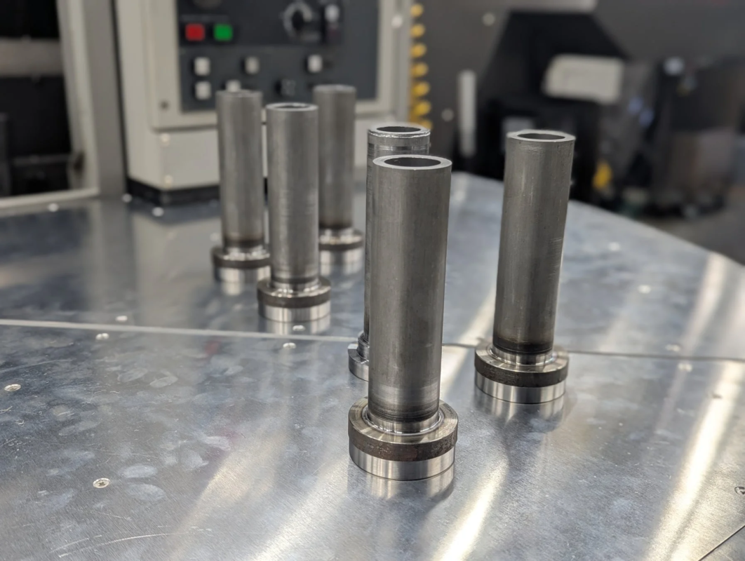 銀色の作業台に、底にフランジが付いた金属製の円筒形部品が複数並べられています。