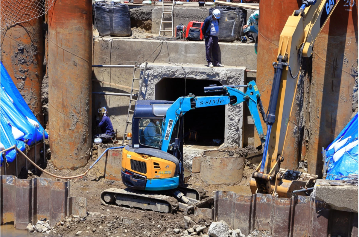 建設現場で、青と黄色のミニショベルカーが活動中。作業員が複数おり、大きな円筒形の構造物やコンクリート製のトンネル入り口が見える。活発な土木工事の様子を捉えた写真。