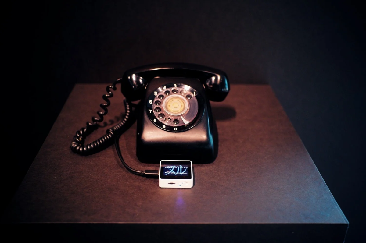 暗い背景のテーブルに置かれた黒いロータリー式の電話機と、その横にケーブルで接続された「文ル」と表示された小型の白いデバイスが写っています。レトロな電話と現代的なガジェットが並ぶ対比的な情景です。