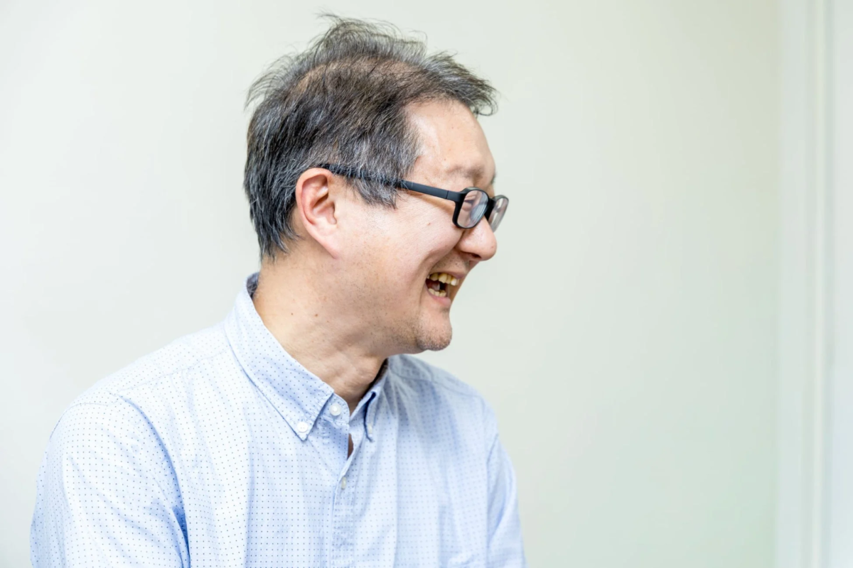 メガネをかけた中高年男性が、白い背景の前で横向きに笑っている様子。水玉模様の青いシャツを着ており、明るく楽しそうな表情を見せています。