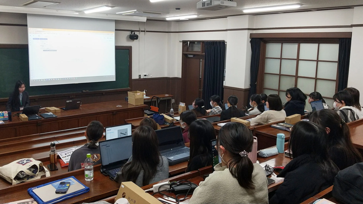 大学の教室で、講師がプロジェクターを使ってプレゼンテーションを行っており、多くの学生がノートパソコンを使用しながら熱心に講義を受けている様子。実践的な学習セッションのようだ。