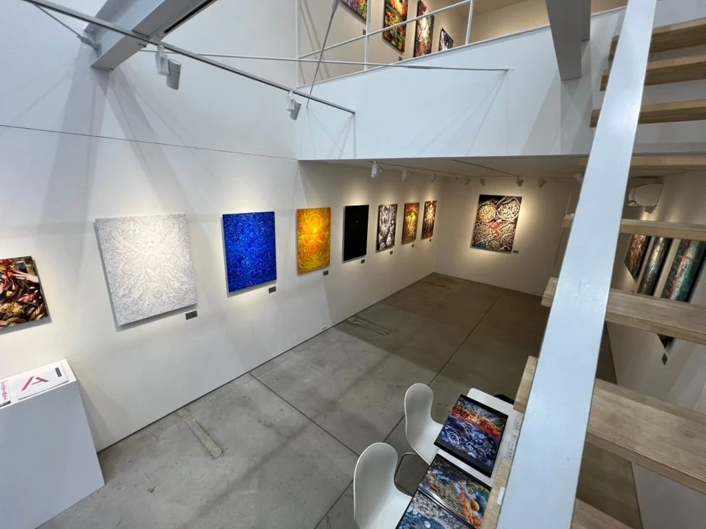 白い壁に鮮やかな絵画が多数展示されたモダンなアートギャラリー。コンクリートの床とシンプルな階段があり、ミニマルな空間で作品が際立っています。