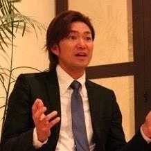 スーツを着た男性が、両手を広げて何かを熱心に説明しているか、スピーチをしている場面