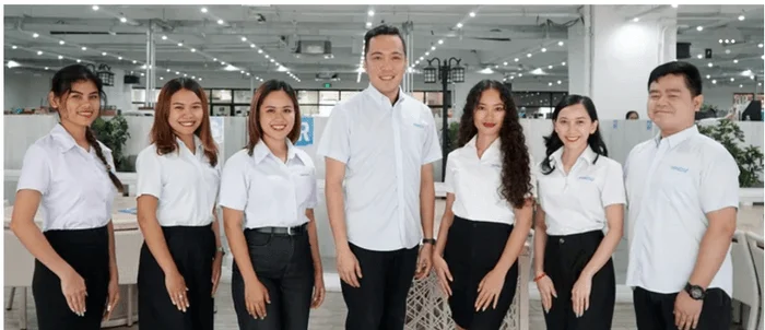 明るいオフィスで白いシャツと黒いボトムスの制服を着た7人の男女が笑顔でカメラに向かって立っている集合写真