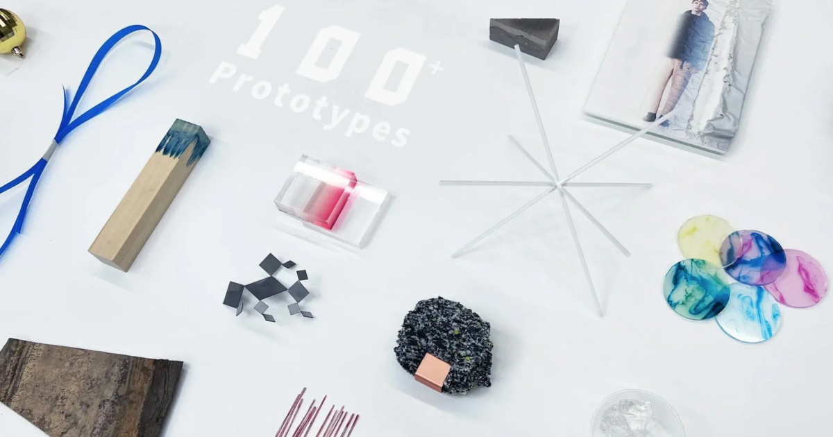 「100 Prototypes」の文字とともに、木材、石、銅、透明樹脂、カラフルなアクリル円盤など、多様な素材や試作品が白い机上に広がる様子。デザインや素材研究、創造的なプロセスを想起させる画像です。