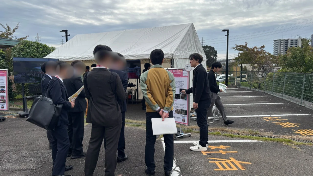 屋外の会場で、複数の人々がVR/AR技術を活用した展示を見学しています。白いテントの前で、参加者たちがディスプレイや説明パネルに注目しており、活発な情報交換が行われている様子です。