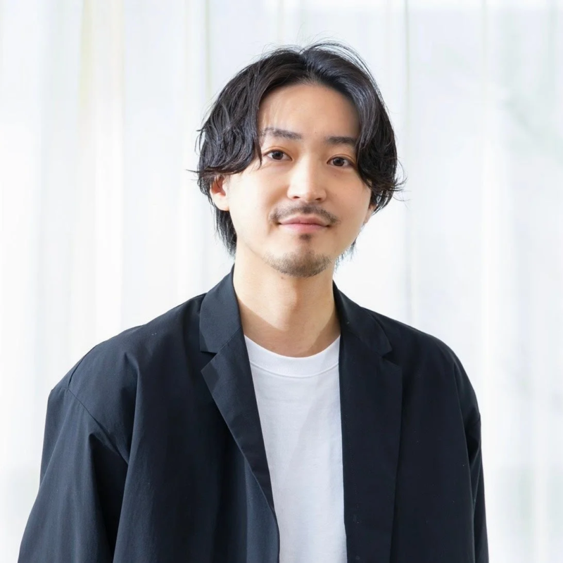 黒いジャケットと白いTシャツを着たアジア人男性が、カメラに向かって優しく微笑んでいるポートレート写真