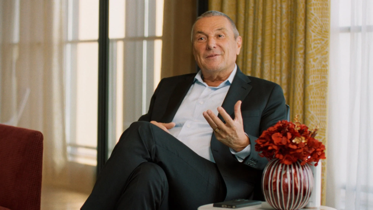 スーツを着た年配の男性が、室内で椅子に座り、身振り手振りを交えながら話している様子。